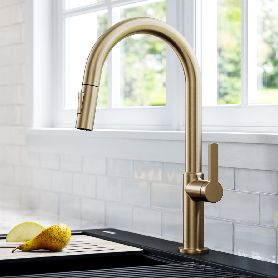 Brushed gold pull-down kitchen faucet with a single handle installed over a black kitchen sink with a wooden cutting board and pear slices.