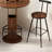 This Walnut Wood Three-Piece Bar Table And Chair Set Features A Round Table With Two Stools And Metal Footrests. It's Compact, Versatile, And Suitable For Small Spaces Such As Dining Rooms, Bars, And Living Rooms.