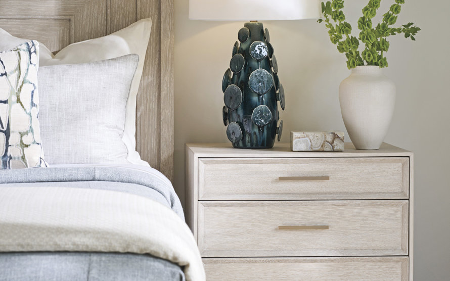 Light wood nightstand with two drawers and gold hardware next to a bed featuring a textured lamp, a stone accent box, and a white vase with greenery.