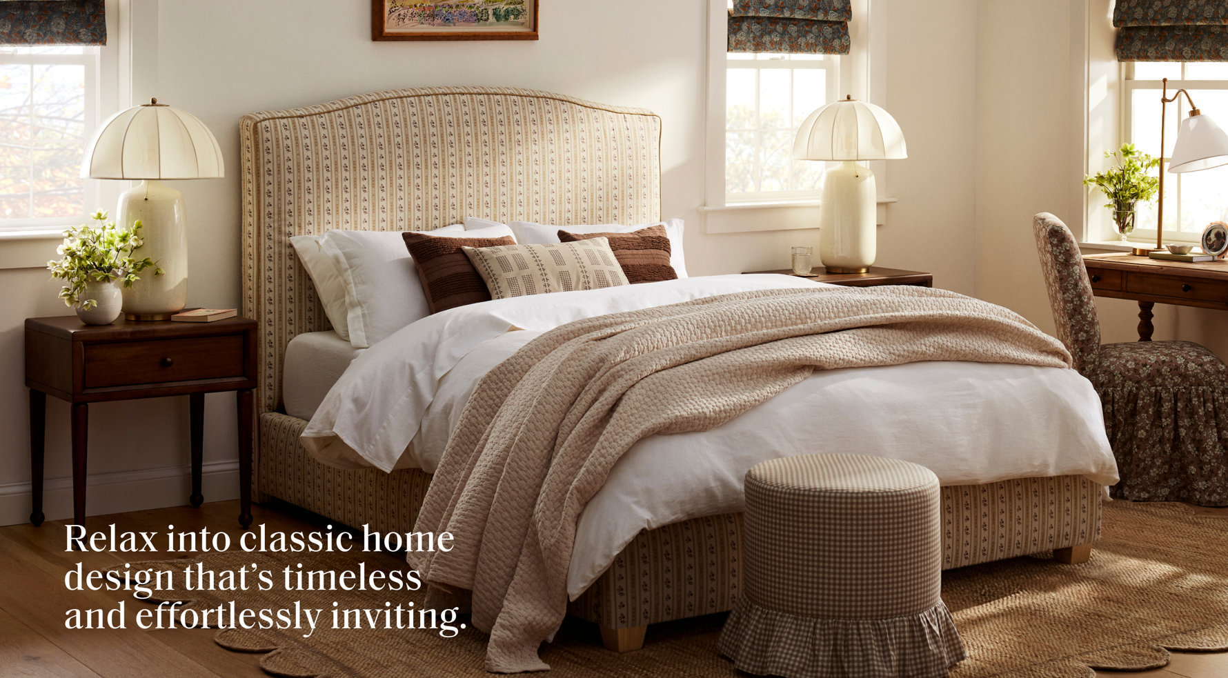 Relax into classic home design that’s timeless and effortlessly inviting. A bedroom featuring a patterned upholstered bed, neutral bedding, two bedside lamps, and a skirted ottoman on a natural fiber rug.