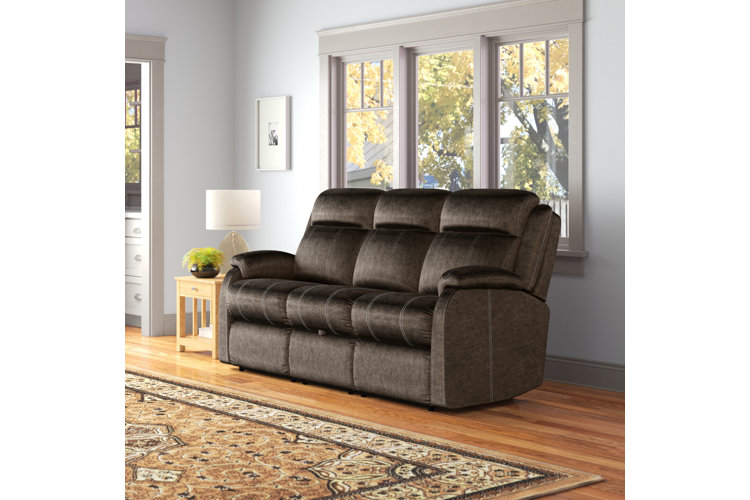A living room with a dark brown suede leather reclining sofa up against a wall with three windows. 