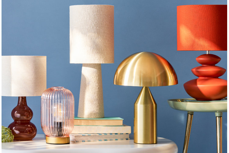 A variety of stylish table lamps arranged on surfaces against a blue background. From left to right: a small brown glass lamp with a white shade, a transparent ribbed glass lantern-style lamp with a gold base, a tall beige fabric-textured lamp sitting on stacked books, a sleek metallic gold mushroom-shaped lamp, and an orange lamp with a stacked-geometric base and matching orange shade resting on a small side table.