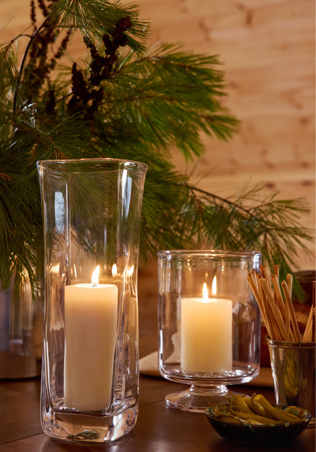 Two glass hurricane candle holders with lit white pillar candles arranged on a wooden table with pine branches, breadsticks, and a bowl of olives.