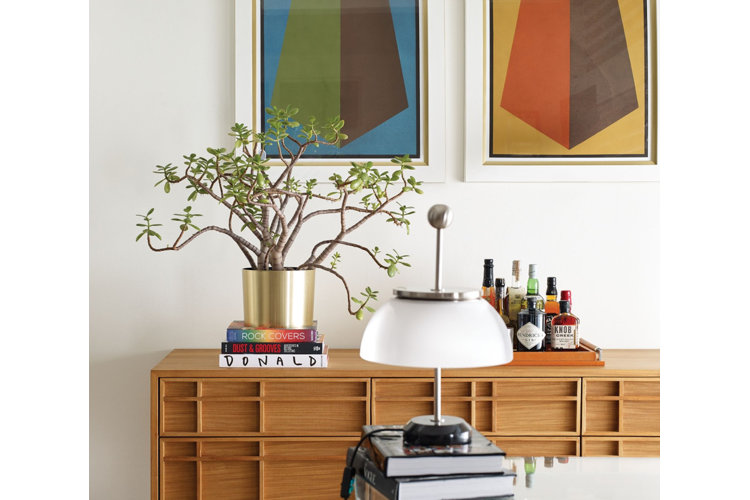A blonde wood dresser with an etched pattern. Atop the dresser, there is a gold metallic planter holding a small tree, three books and a decorative tray holding tiny bottles. On the wall, there are two brightly-coloured geometric paintings. The room is light and bright.