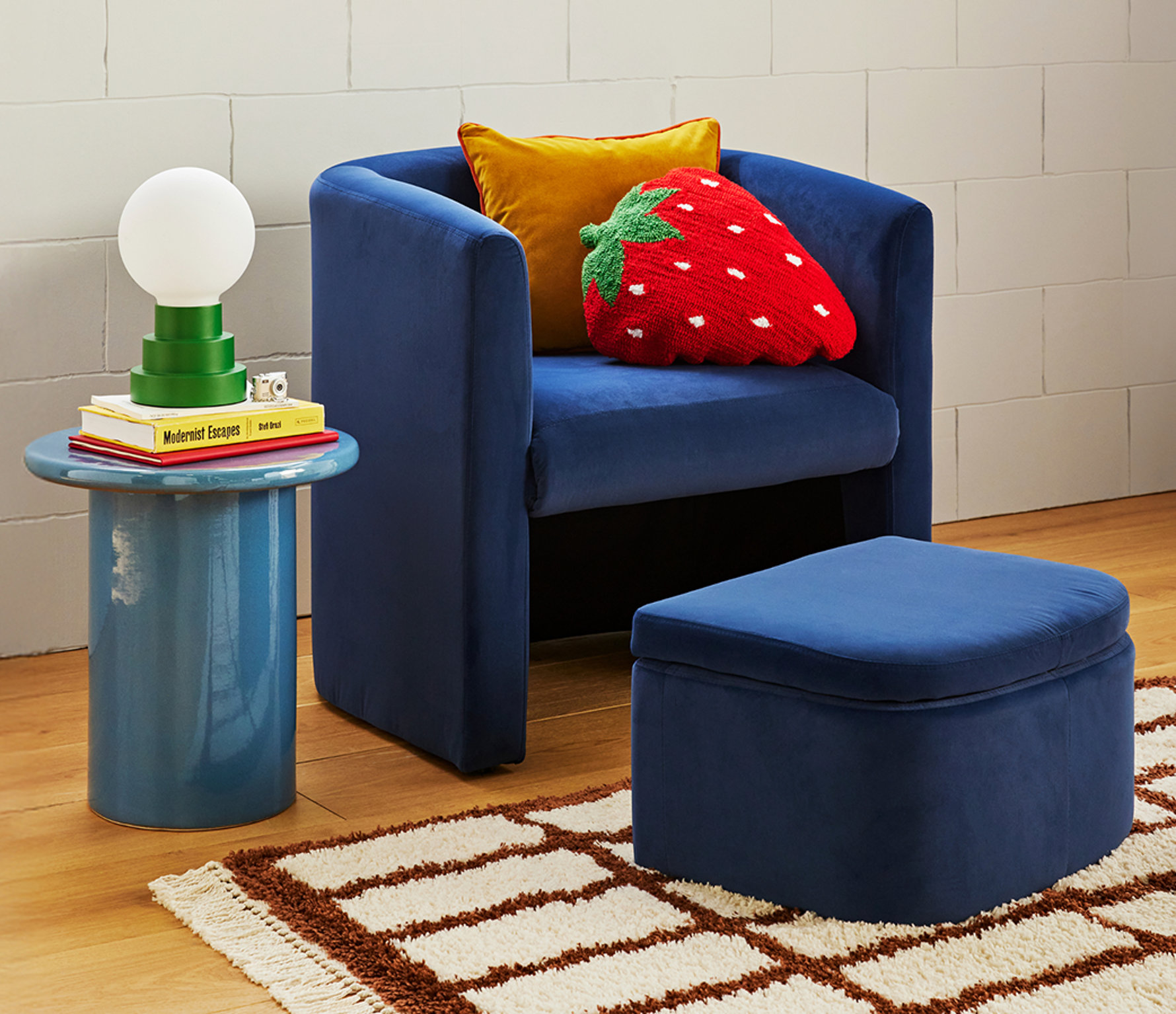 A navy blue velvet barrel chair and matching ottoman styled with a bright red strawberry throw pillow.