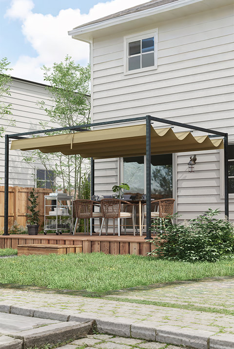 Black metal pergola with a retractable beige canopy set over a wooden patio deck with a dining table and woven chairs next to a white house.