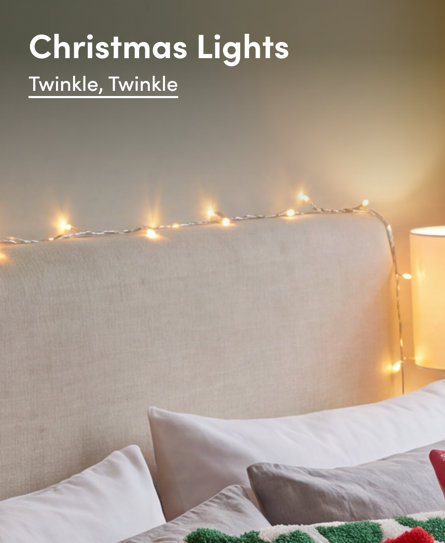 Christmas Lights, Twinkle, Twinkle. Warm fairy lights draped over a beige upholstered headboard in a cosy bedroom setting.