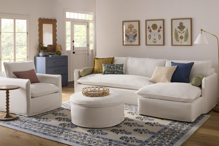 traditional style living room with a white sofa matching accent chair and matching drum shaped ottoman with white upholstery blue and white traditional style area rug and wooden traditional end table a wicker tray on the ottoman and floral wall art behing the sofa