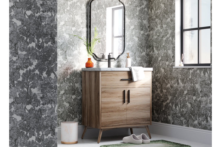 Powder room with black and white toile wallpaper a light wood midcentury modern bathroom vanity with an oval black frame vanity mirror an orange vase with green plant a small white and gold waste basket and green bath mat on the floor with slippers