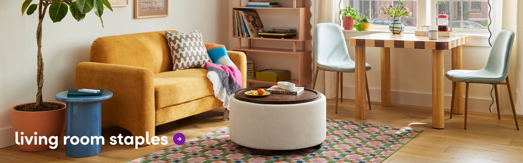 A bright living room featuring a yellow loveseat, multicolor geometric area rug, white boucle ottoman, pink bookshelf, dining table and light blue chairs near a sunny window.