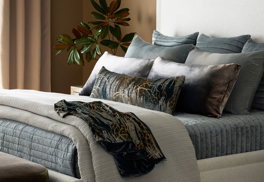 Cozy bedroom scene featuring a bed with textured gray bedding, plush velvet pillows, and a decorative botanical print throw blanket and matching lumbar pillow next to a magnolia plant.