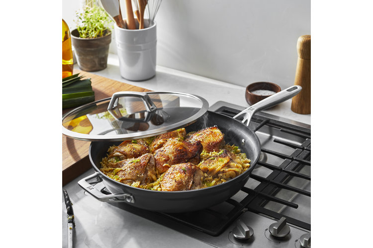 Lidded frying pan with cooked chicken on a gas stove with continuous grates.