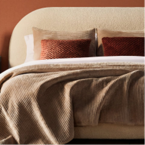 Modern arched headboard in cream bouclé fabric layered with tan and rust-colored decorative pillows and a ribbed beige throw blanket.