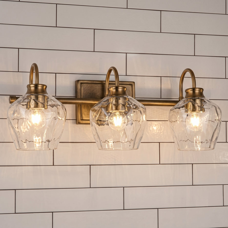 Three-light bathroom vanity fixture with gold-toned hardware, clear hammered glass shades, and a black mounting plate installed on a white subway tile wall above a mirror.
