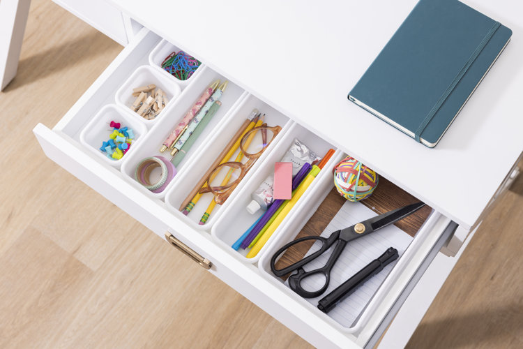 An open desk drawer filled with office supplies in organized bins, including small containers for thumb tacks and paper clips.