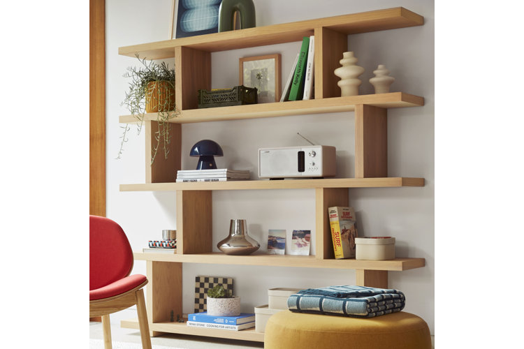 A modern living room sitting area featuring a low-profile red upholstered accent chair with light wood legs, positioned on a textured off-white area rug next to a white ceramic pedestal side table. To the right, a round yellow pouf ottoman holds a folded blue patterned blanket. In the background, a large, asymmetrical wooden bookshelf displays various books, small plants, decor items, and a small blue mushroom-shaped lamp, while a sliding glass door on the left reveals tall cacti growing outside.