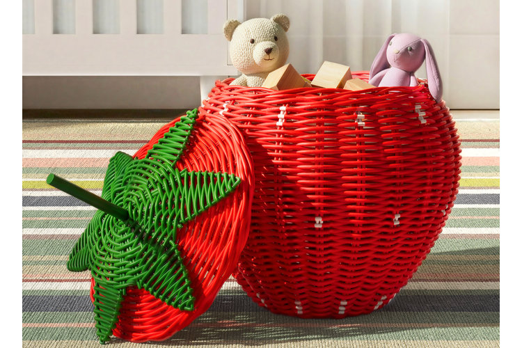 A bright red strawberry-shaped woven wicker storage basket with a green leaf lid, filled with stuffed animals and wooden blocks on a striped rug