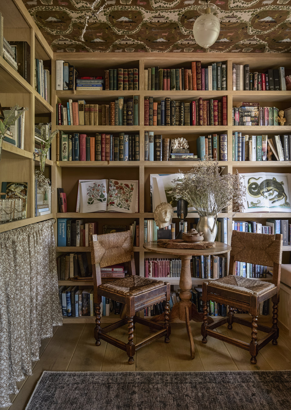 Bibliothèque de maison chaleureuse avec une étagère en bois intégrée remplie de livres, une petite table d'appoint ronde à piédestal en bois, deux chaises rustiques en bois à assise de jonc, ainsi que des éléments décoratifs dont un motif floral au plafond et un arrangement de fleurs séchées.
