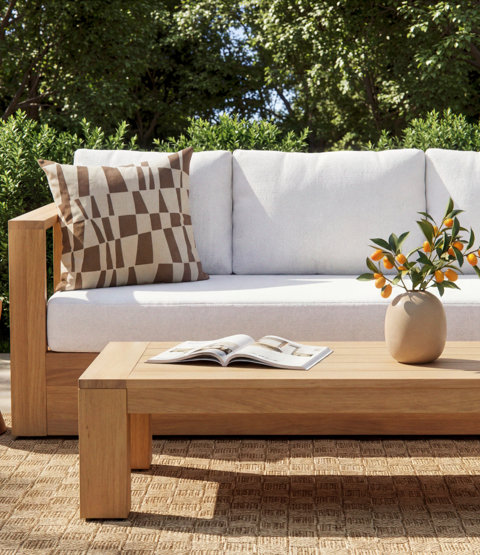 Modern outdoor sofa with teak frame, white cushions, geometric patterned throw pillow, and a coffee table decorated with a vase of kumquats on a woven patio rug.