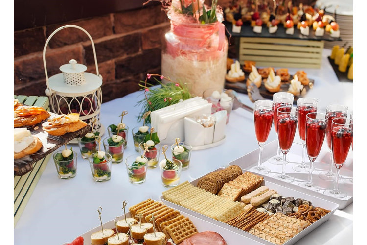 A table with a bunch of snacks and single serve appetizers beside a tray of red cocktails in champagne flutes.