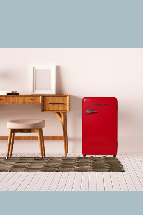 A red retro-style mini fridge sits on a white wood floor next to a mid-century modern wooden desk with a matching stool and an empty picture frame.