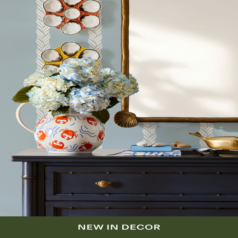 Coastal decor vignette featuring a white pitcher with orange crab and blue wave pattern holding blue hydrangeas, displayed on a navy bamboo dresser with brass shell mirror, colorful oyster plates on the wall, and brass whale figurine. Text reads 'NEW IN DECOR'.