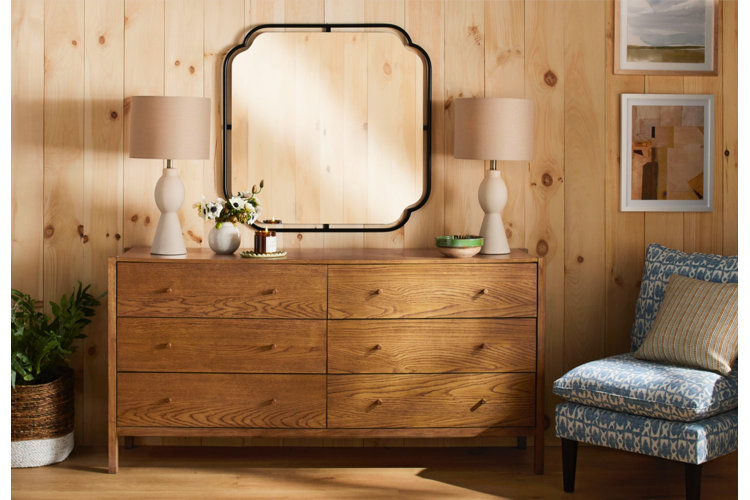 A rustic modern lakehouse bedroom vignette featuring a wide oak dresser against light pine wood wall paneling, styled with a square black-framed mirror, two matching beige table lamps, and a blue pattern-print accent chair