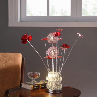 Lampe de table moderne en aluminium chromé, composée de 4 roses rouges à 6 pétales et de 2 boutons de fleurs rouges et argentés, sur un vase hexagonal carré. Idéale pour le salon ou comme lampe de chevet. Lampe LED.