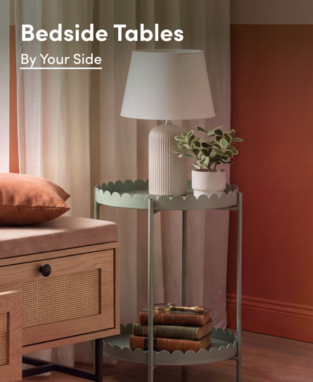 Bedside Tables: By Your Side. A sage green scalloped metal side table styled with a lamp, a small potted plant, and books next to a rattan bed bench in a warm-toned bedroom.