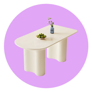Off-white dining table with a smooth oval top and two fluted, pedestal-style bases, styled with a small blue glass vase and a bowl of limes against a purple circular background.