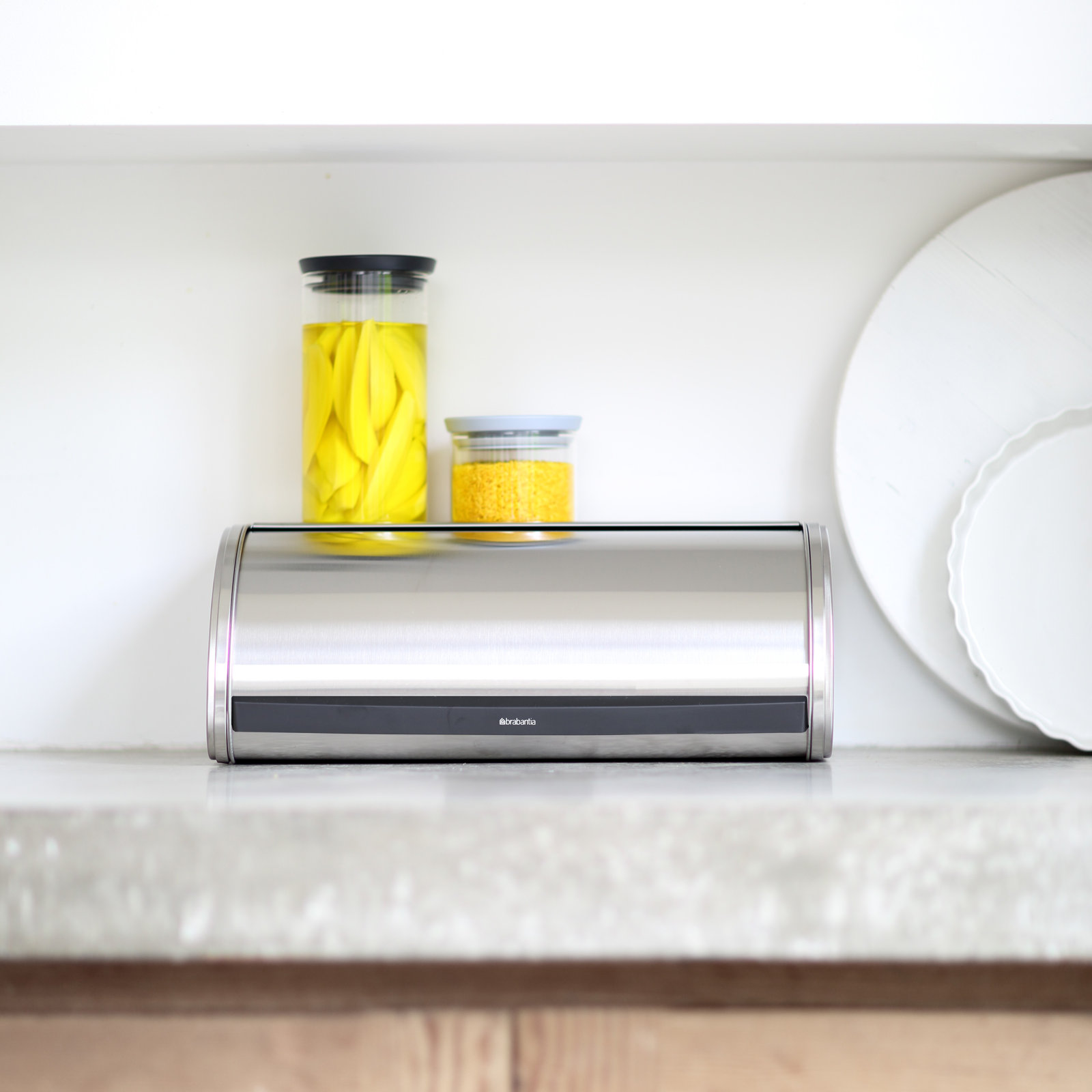 Brushed stainless steel roll-top bread box on a kitchen counter with canisters on its flat top.