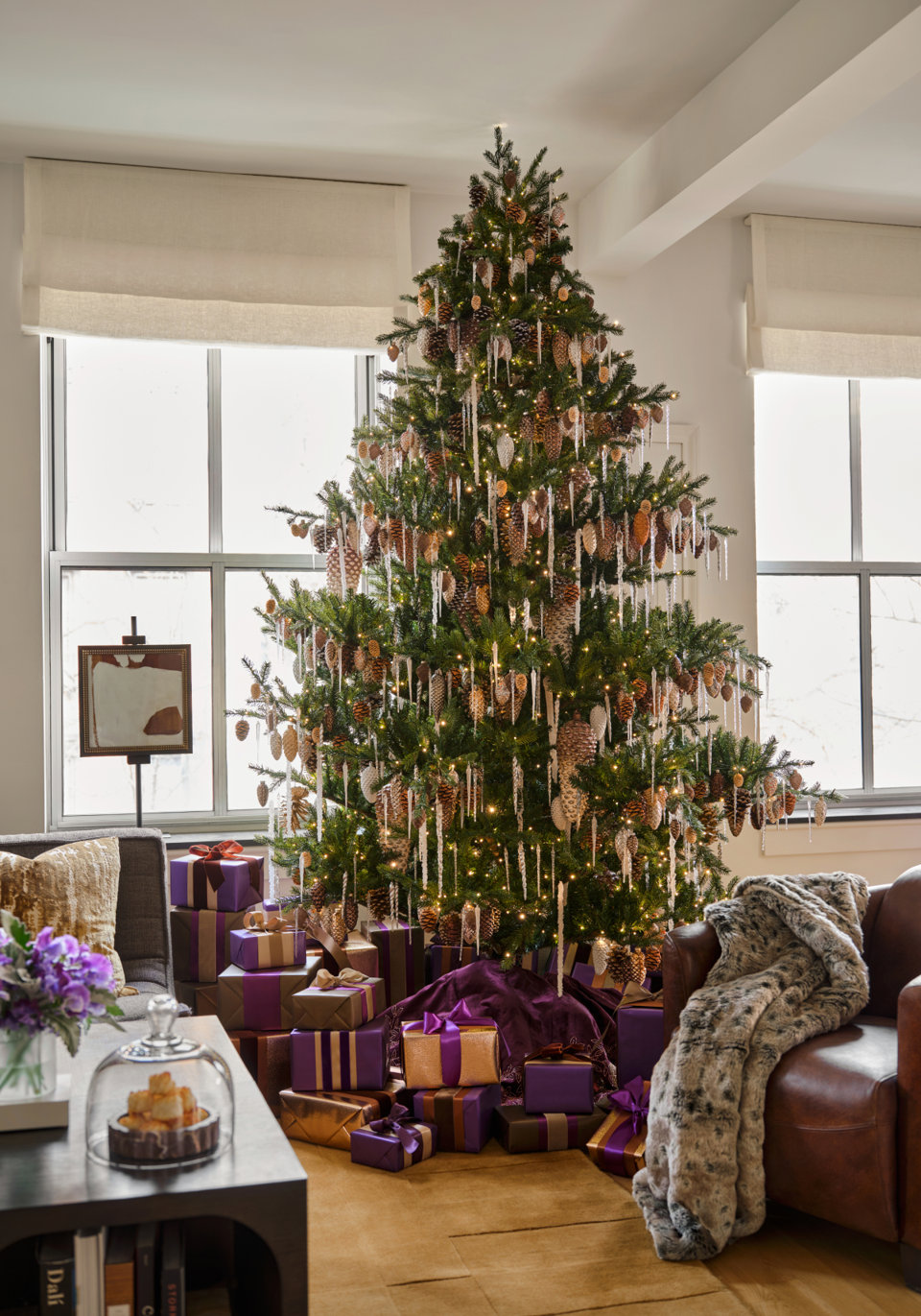 A Christmas tree decorated with pinecones and icicle ornaments stands in a living room, surrounded by purple and gold wrapped gifts.