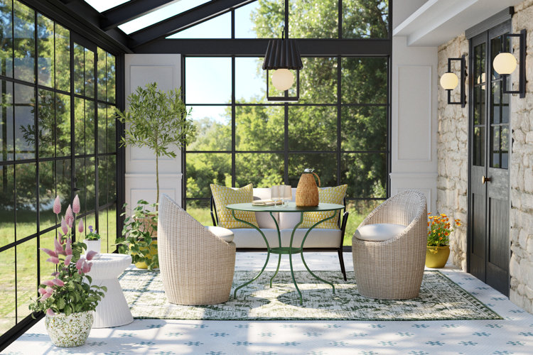 Woven wicker chairs and small round table in a bright sunroom with black-framed windows, patterned rug, and lush greenery views.