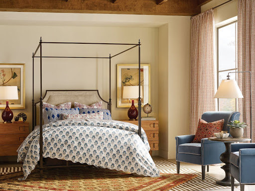 A bedroom with bright patterns: an upholstered bed layered with patterned blankets, a patterned rug, and two armchairs in muted blue.