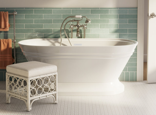 White freestanding bathtub with a vintage-style faucet positioned against a sage green subway tile wall, featuring a decorative white rattan stool in the foreground.