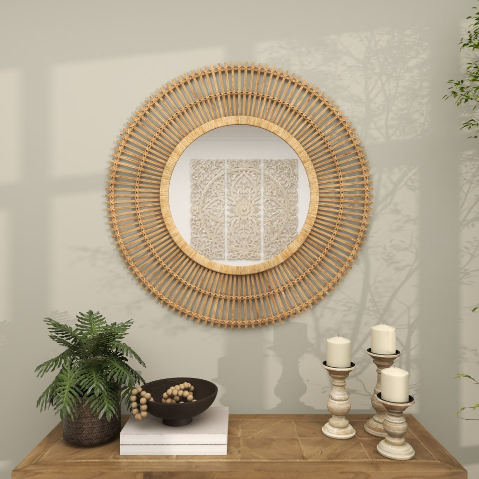 Round wall mirror with a decorative natural wood sunburst frame hanging above a console table styled with a potted plant, a bowl of wooden beads, and three pillar candles.