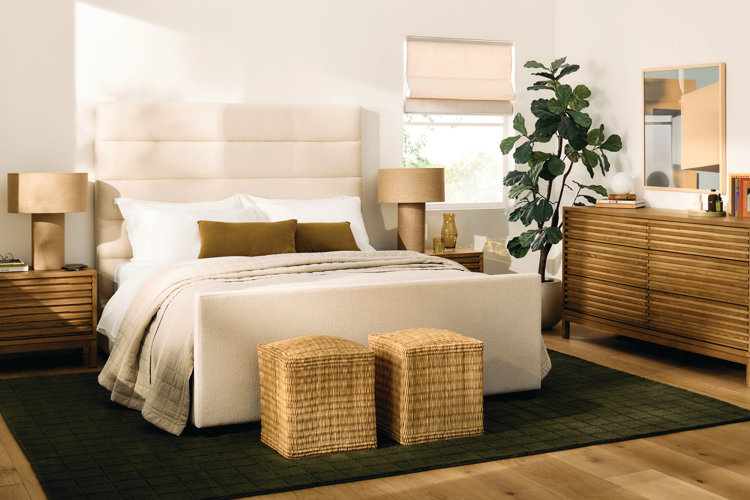 A modern bedroom filled with warm light and natural textures. The center features a cream upholstered bed with a tall horizontal-channel headboard, made with white linens, a tan quilted blanket, and olive green accent pillows. At the foot of the bed sit two square woven rattan ottomans on a dark forest green grid-patterned rug.