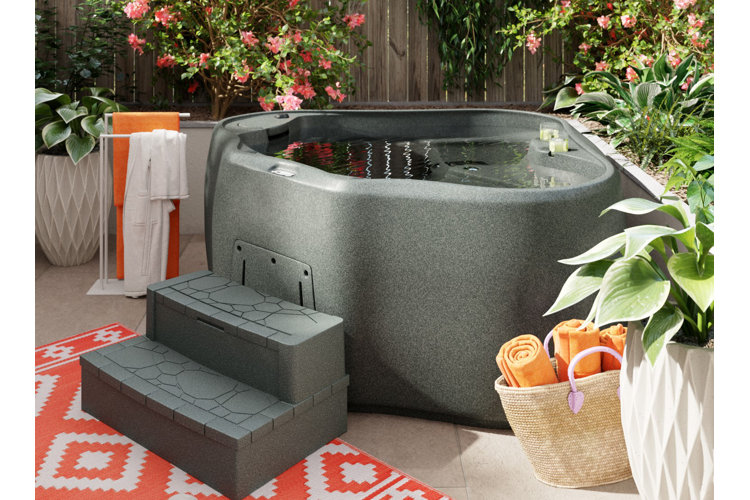 Gray stone-look corner hot tub with steps and surrounded by a plant border and a basket of towels.