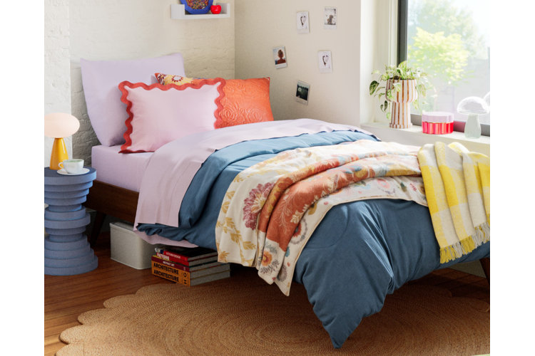 Bed with an array of colorful bedding including a blue divet, pink sheets, floral quilt, and a yellow throw blanket. Blue nightstand with a yellow lamp