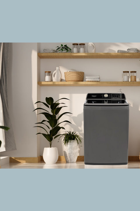 A gray top-load washing machine sits in a bright, modern laundry room with wooden floating shelves filled with baskets, containers, and plants.