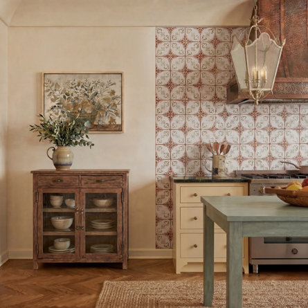 European farmhouse-style kitchen featuring a decorative tile backsplash, wooden cabinet displaying dishware, a pitcher with greenery, pendant lantern lighting, and a rustic green island table