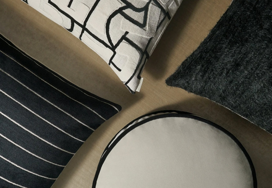 Top-down view of four decorative throw pillows in varying textures and patterns, including a black and white geometric print, a black-and-white striped rectangular pillow, a circular cream pillow with black piping, and a textured black faux fur pillow on a neutral background.
