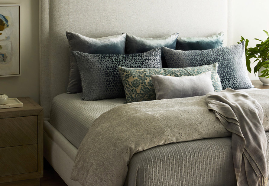 Close-up of a neatly made bed featuring a light beige upholstered headboard, textured gray bedding, and a layered collection of velvet and patterned decorative pillows in shades of blue and cream.