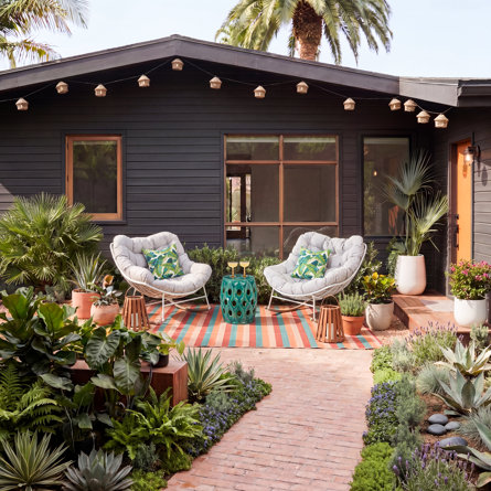 Terrasse extérieure avec deux chaises berçantes grises, une table d'appoint en céramique turquoise et un tapis rayé coloré devant une maison aux murs sombres.