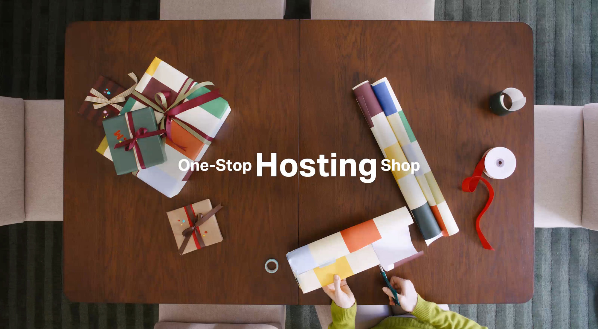 One-Stop Hosting Shop with a top-down view of a person wrapping colorful gifts on a wooden table.