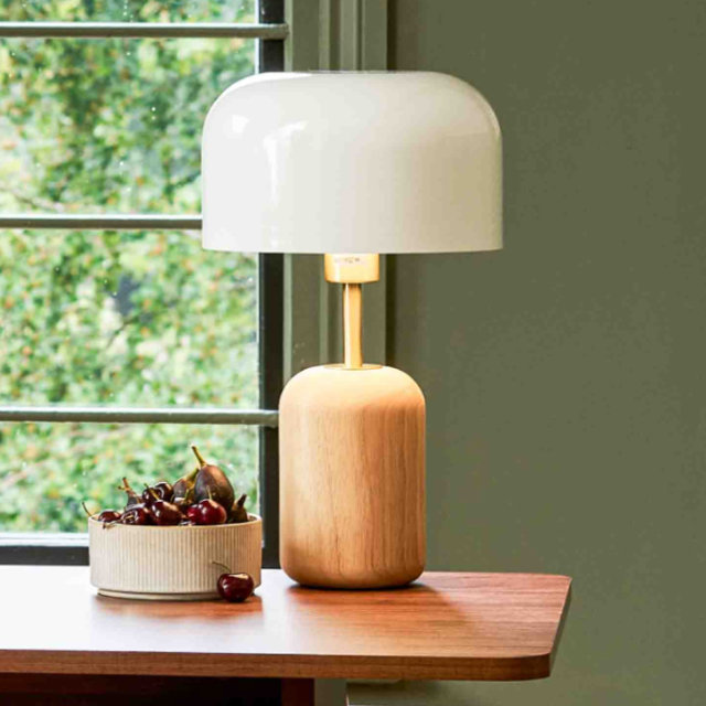 Modern table lamp with a natural wood base, brass neck, and large white dome shade sitting on a wooden desk next to a bowl of cherries.