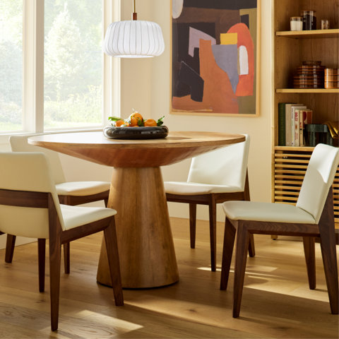 Mid-century modern dining room featuring a round wooden pedestal table surrounded by four chairs with cream upholstery and dark wood frames, lit by a white ribbed pendant lamp.