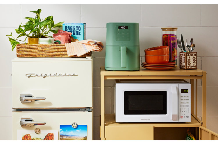 A cozy dorm kitchen setup featuring a retro ivory mini fridge styled with a woven tray, potted plant, and snacks on top. Next to it, a yellow utility cart holds a sage green air fryer, stacked orange bowls, silverware, and a compact white countertop microwave. 
