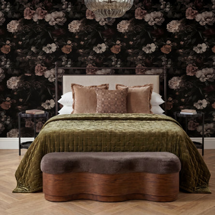 A romantic moody bedroom featuring dark floral wallpaper, an upholstered bed frame with an olive green velvet quilt and blush velvet pillows, round side tables, and an elegant crystal chandelier