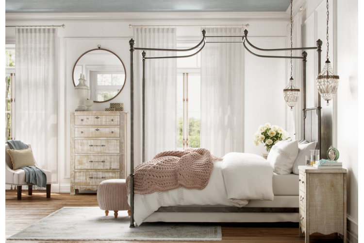 French country bedroom with white sheer curtains, a black metal canopy bed, and distressed wood furniture.